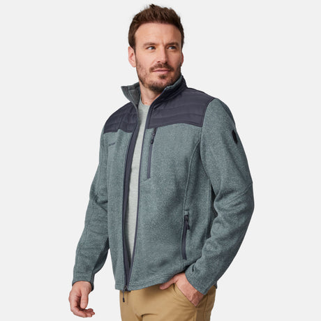 A man with short brown hair and a beard wears the Free Country Men's Frore Knit Fleece Jacket in light gray with dark gray shoulder panels, a convertible collar, a light gray T-shirt, and tan pants. He looks to the side with one hand in his pocket.