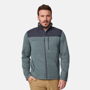 A man wears the Free Country Men's Frore Knit Fleece Jacket in light gray with a dark gray quilted shoulder overlay and chest pocket, paired with khaki pants, standing against a plain white background.