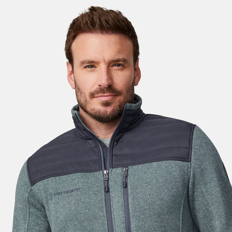 A man with short brown hair and a trimmed beard is wearing the Free Country Men's Frore Knit Fleece Jacket, featuring dark gray shoulder panels and a convertible collar, standing against a plain white background.