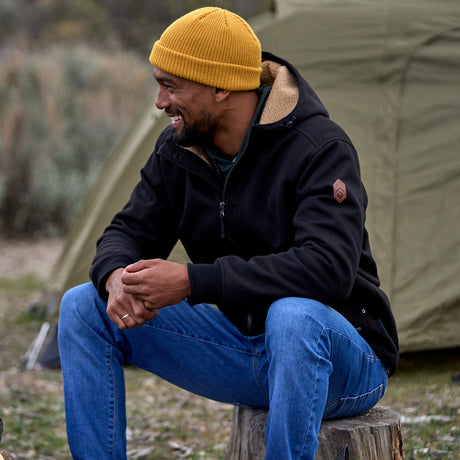 Men’s Free Country black hooded jacket featuring full front zipper, two side pockets, and zippered chest pocket. Includes utility patch on left sleeve and subtle brand label at lower hem. Styled with blue jeans for a laid-back cold-weather look highlighting functional design and casual edge.