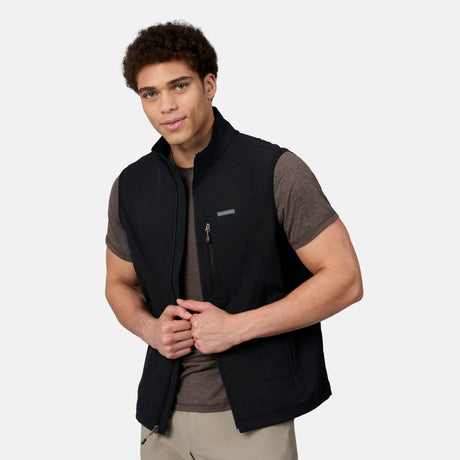 A young man with curly hair wears the Free Country Men's Stretch Rip Stop Adventure Vest in black, layered over a brown t-shirt and beige pants. He faces forward, holding the open sides of the vest against a plain light background.