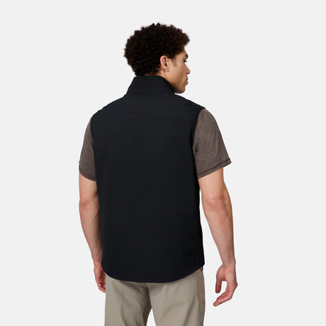 A man faces away, wearing the Free Country Men's Stretch Rip Stop Adventure Vest in black over a brown short-sleeve shirt and beige pants, set against a plain light gray background.