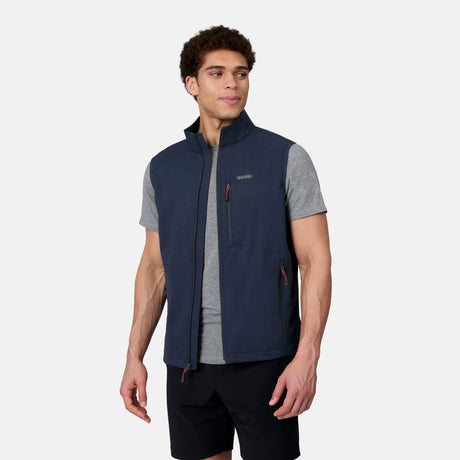 A man in a Free Country Men's Stretch Rip Stop Adventure Vest—navy, lightweight, wind resistant, and quick-drying—over a light gray tee and black shorts stands turned slightly to the side against a plain light background.
