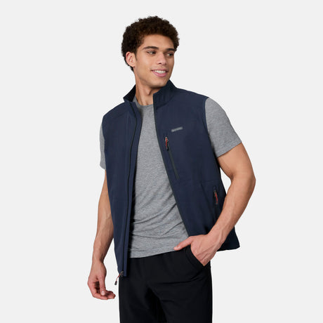 A man wearing a gray t-shirt and the Free Country Men's Stretch Rip Stop Adventure Vest in navy blue stands with his hand in his pocket, looking to the side against a plain light background.