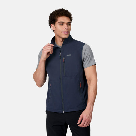 A man stands against a plain light background, looking to the side and holding the collar of his Free Country Men's Stretch Rip Stop Adventure Vest, worn over a gray short-sleeve t-shirt.