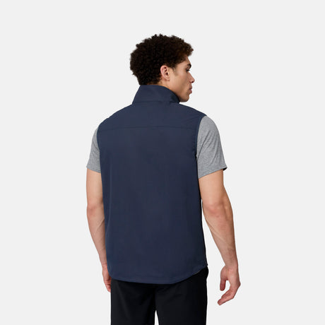 A man with short curly hair is seen from behind wearing the Free Country Men's Stretch Rip Stop Adventure Vest in navy over a gray short-sleeve shirt and black pants, against a plain light gray background.