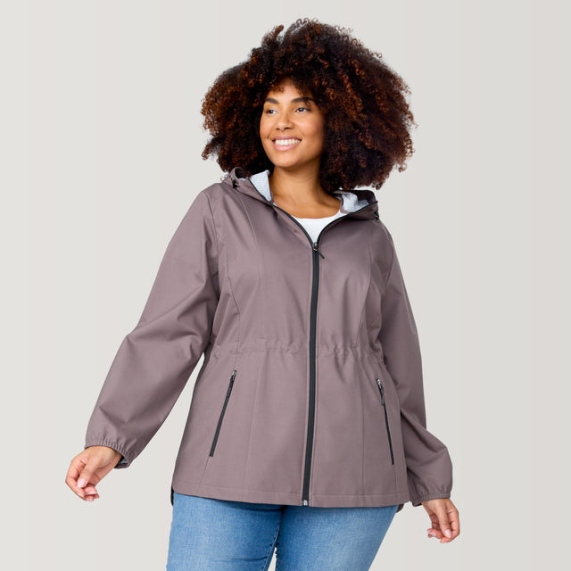 A woman with curly hair smiles and looks to the side, wearing the Free Country Women's Plus Size X2O Anorak Rain Jacket in taupe over a white top and blue jeans, set against a plain light background.