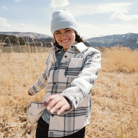 Person wearing a plaid jacket and beanie standing in a field with mountains in the background