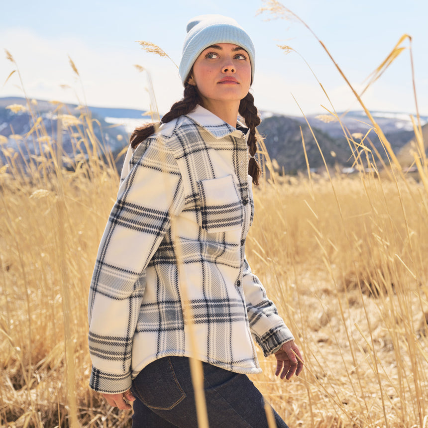 Woman in a plaid jacket and beanie standing in a field with mountains in the background