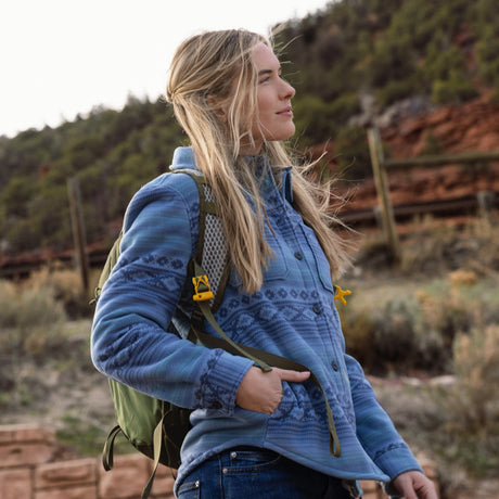 A woman hiking outdoors wearing a blue patterned button-up fleece jacket, blue jeans, and a green backpack, with a blurred natural landscape in the background.