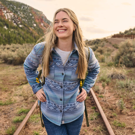 A woman smiling outdoors on railroad tracks, wearing a blue and gray patterned button-up shirt over a white undershirt with a backpack visible.