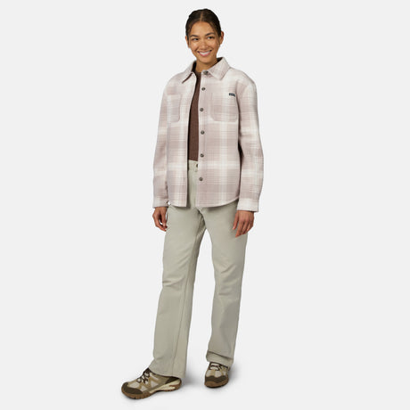 [Model is wearing size S.] A woman modeling a light pink and beige plaid button-up shirt jacket over a dark brown shirt, paired with light gray pants and brown hiking shoes, against a plain light gray background.