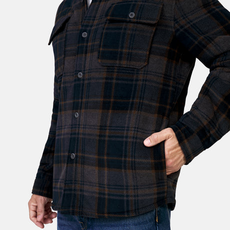A man models the Free Country Men's Chill Out Fleece Shirt Jacket, featuring a dark plaid pattern, sherpa lining, two chest pockets, and side pockets, styled with blue jeans against a white background.