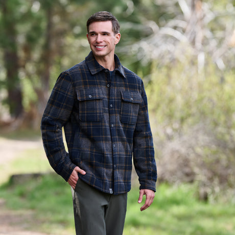 A man stands outdoors on a path, smiling amid trees and greenery, while wearing the Free Country Men’s Chill Out Fleece Shirt Jacket in a blue and brown plaid pattern with sherpa lining.