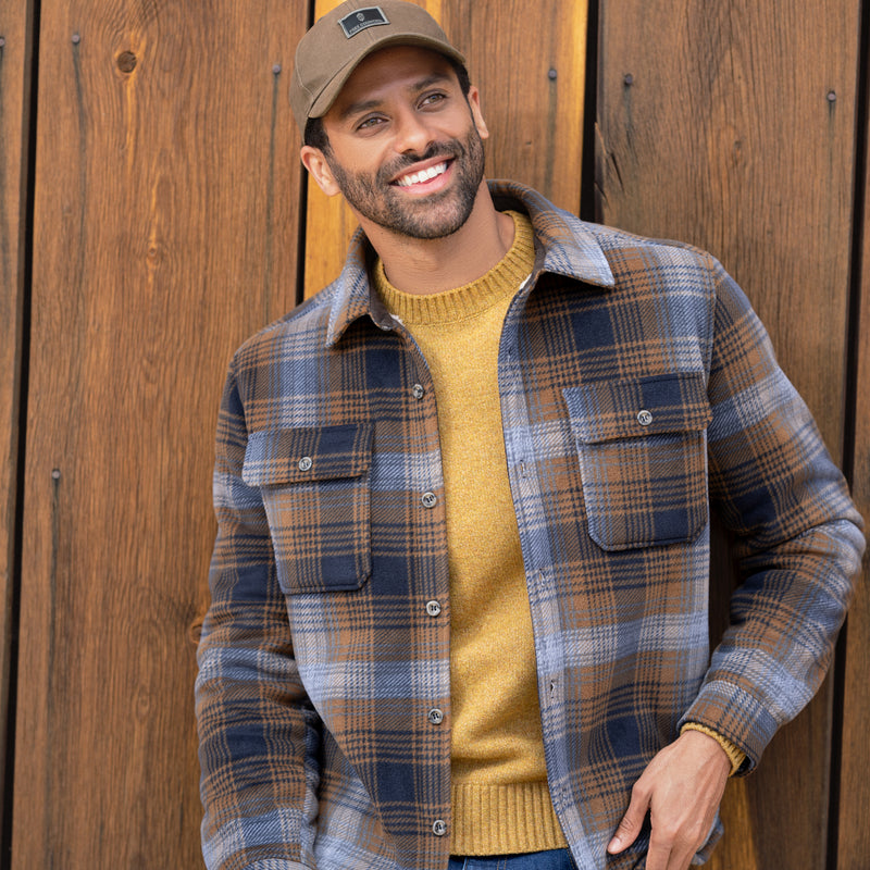 Man wearing a plaid shirt and yellow sweater standing against a wooden background