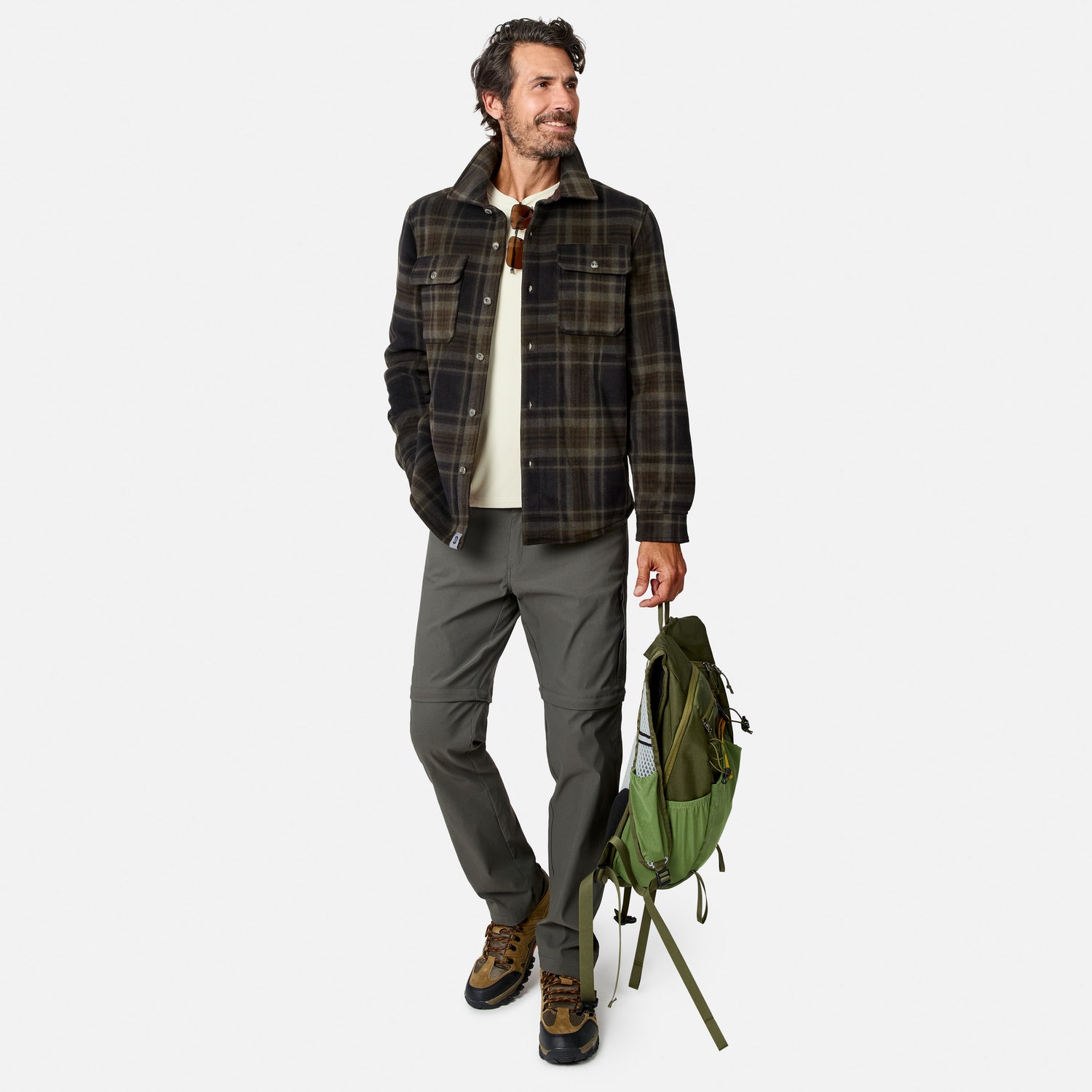 A man wearing the Free Country Men’s Chill Out Fleece Shirt Jacket stands holding a green backpack. He pairs the plaid sherpa-lined shirt-jacket with a cream shirt, gray pants, and hiking boots. With short curly hair and a beard, he smiles while looking to the side.