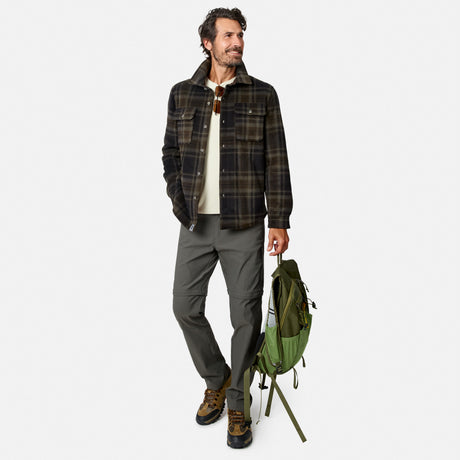 A man wearing the Free Country Men’s Chill Out Fleece Shirt Jacket stands holding a green backpack. He pairs the plaid sherpa-lined shirt-jacket with a cream shirt, gray pants, and hiking boots. With short curly hair and a beard, he smiles while looking to the side.