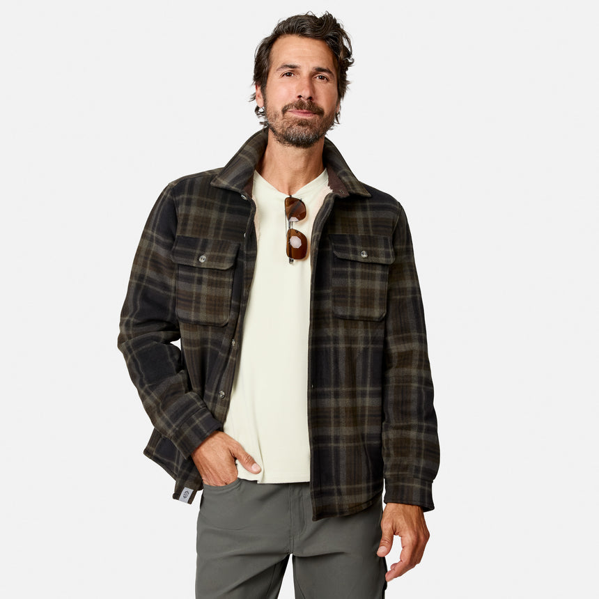 A man with dark hair and a beard is wearing the Free Country Men’s Chill Out Fleece Shirt Jacket in a dark plaid pattern over a white shirt and gray pants, with sunglasses hanging from his shirt collar. He stands facing the camera against a plain white background.