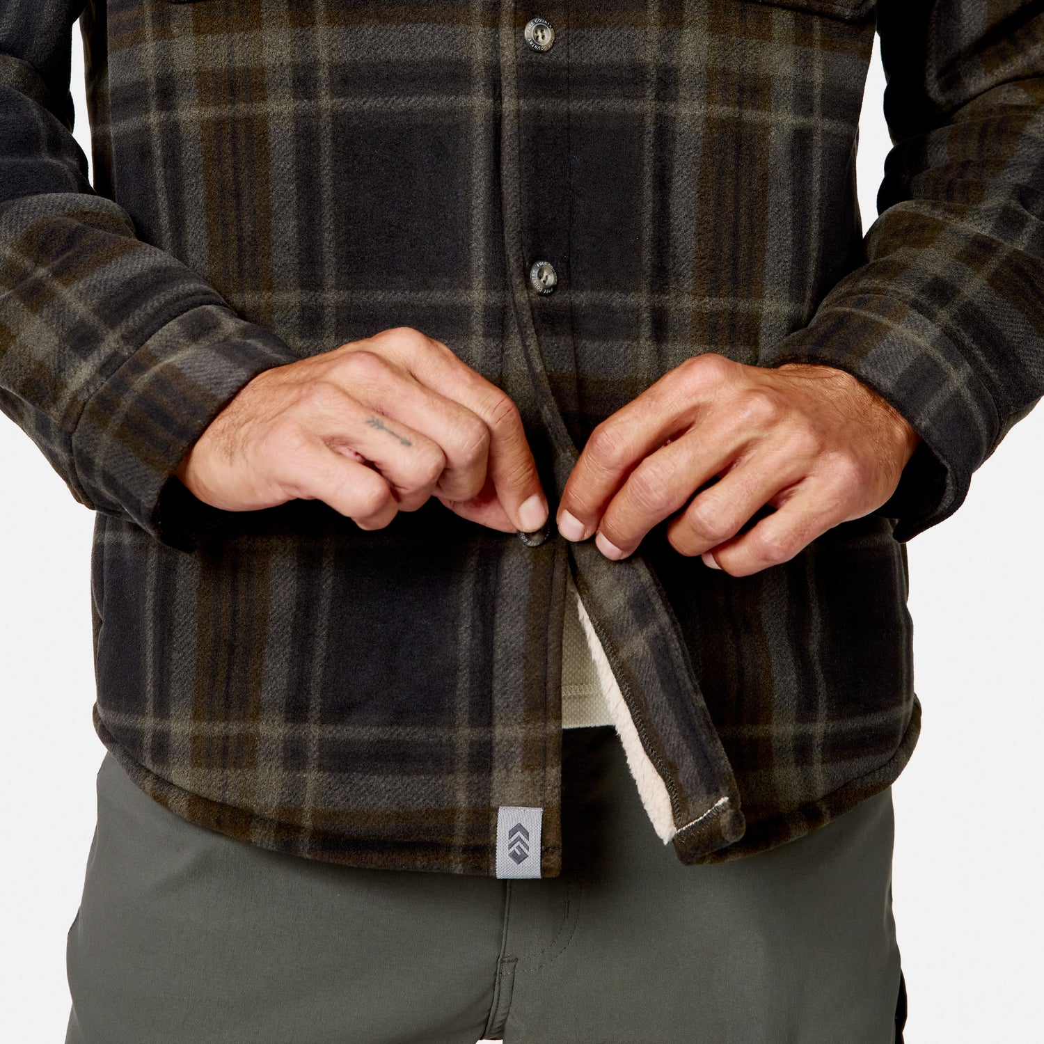 A person wearing the Free Country Men’s Chill Out Fleece Shirt Jacket and gray pants is buttoning up the bottom of the shirt jacket. Only their hands and torso are visible.