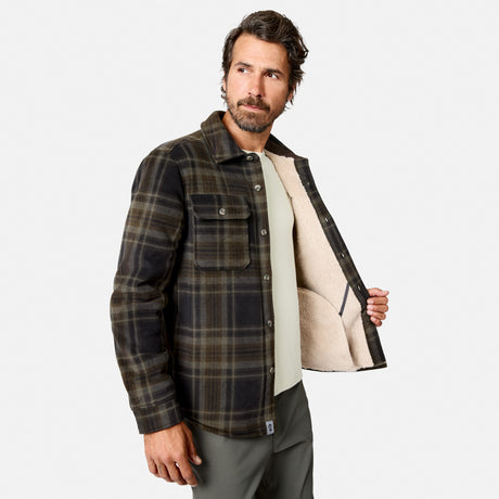 A man with short dark hair and a beard stands facing forward, casually dressed in the Free Country Men’s Chill Out Fleece Shirt Jacket. The green and brown plaid shirt jacket features a light-colored sherpa lining, which he reveals by holding one side open, highlighting the comfortable interior of this outdoor apparel.