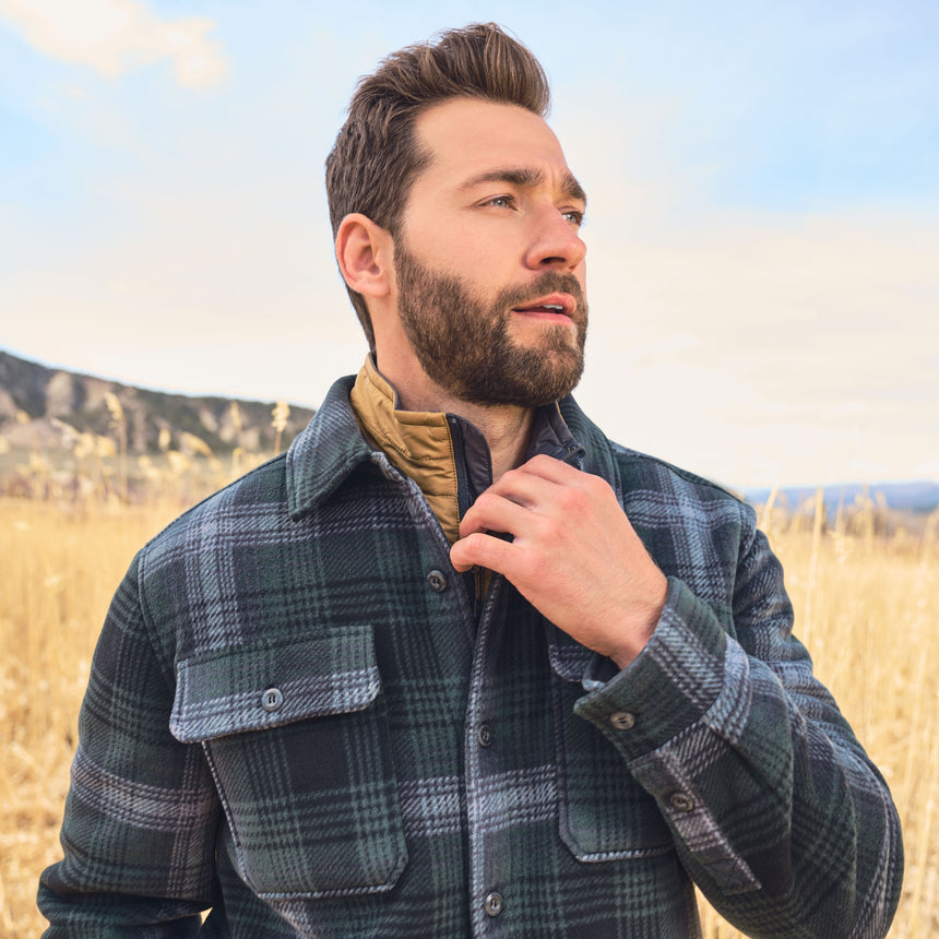 Man wearing a plaid jacket in a field with mountains in the background
