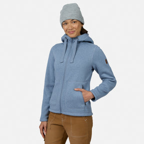 [Model is wearing size S.] A woman wearing a heathered light blue zip-up hooded jacket, a light gray beanie, and brown pants, standing against a plain light gray studio background.
