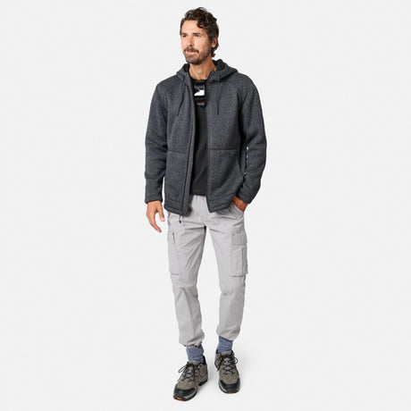 [Model is wearing size M.] A man with short curly hair and a beard stands against a white background wearing the Free Country Men’s Mountain Fleece Jacket in dark colors, paired with a black shirt, light gray cargo pants, blue socks, and gray hiking shoes. He is looking slightly to the left.