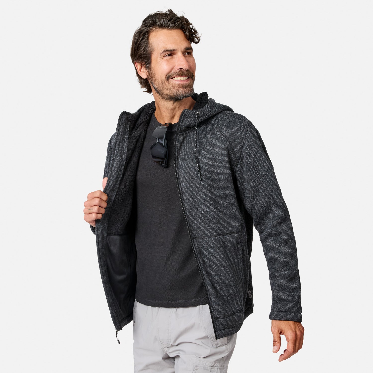 [Model is wearing size M.] A bearded man smiles while wearing a dark Men’s Mountain Fleece Jacket from Free Country, layered over a black shirt and light shorts. He holds the jacket open slightly, with a pair of sunglasses hanging from his shirt collar. The background is plain white.