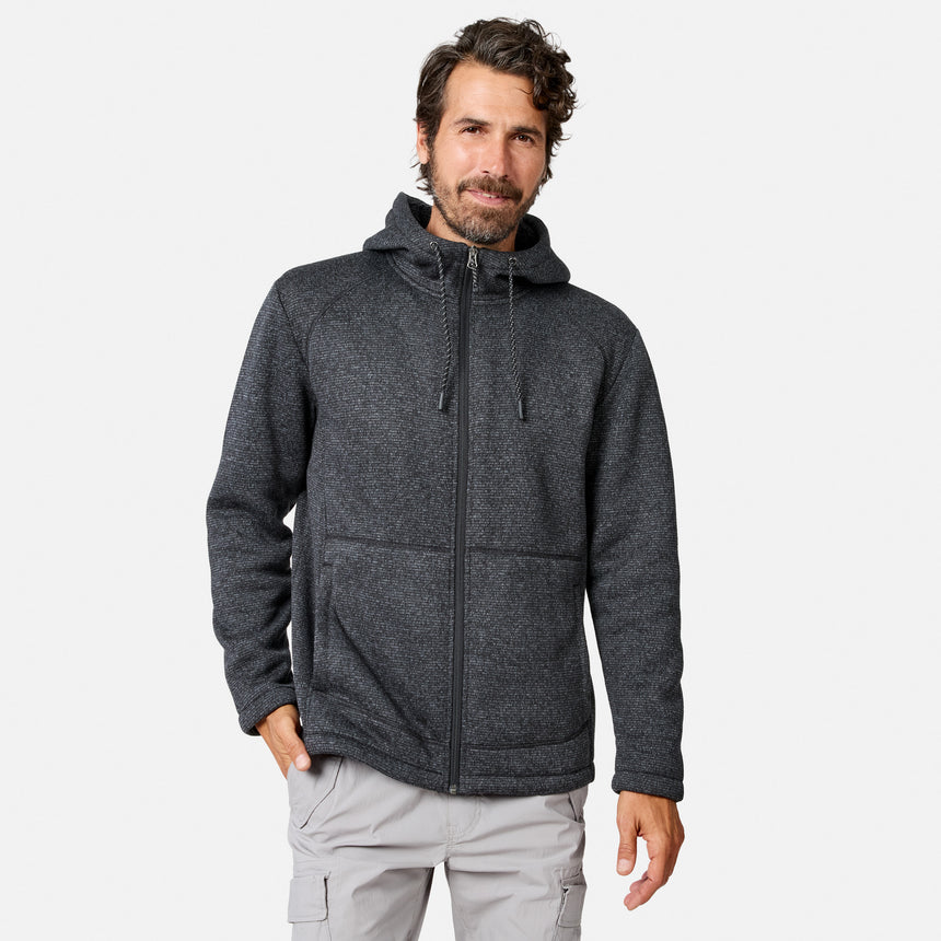 [Model is wearing size M.] A man with wavy brown hair and a beard wears a Men’s Mountain Fleece Jacket by Free Country in Black Texture, known for its lightweight warmth, paired with light gray cargo pants. He stands against a plain white background, with one hand in his pocket.