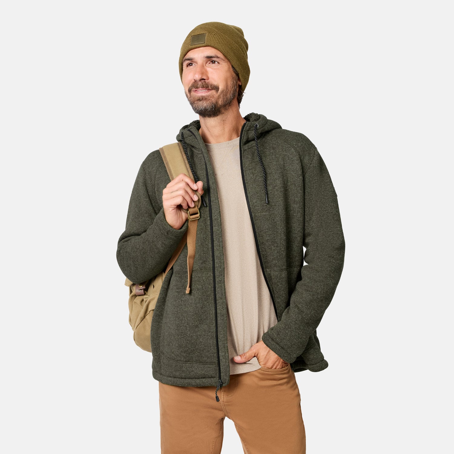 [Model is wearing size M.] A man wearing a green Free Country Men’s Mountain Fleece Jacket, tan beanie, and tan pants stands smiling with a tan backpack over his shoulder against a plain white background.