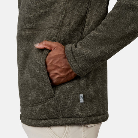[Model is wearing size M.] A man is shown from the side against a plain background, wearing the Free Country Men’s Mountain Fleece Jacket in textured dark green. Designed for lightweight warmth, this mountain fleece-style jacket features a small cloth tag near the front pocket, where his right hand rests.