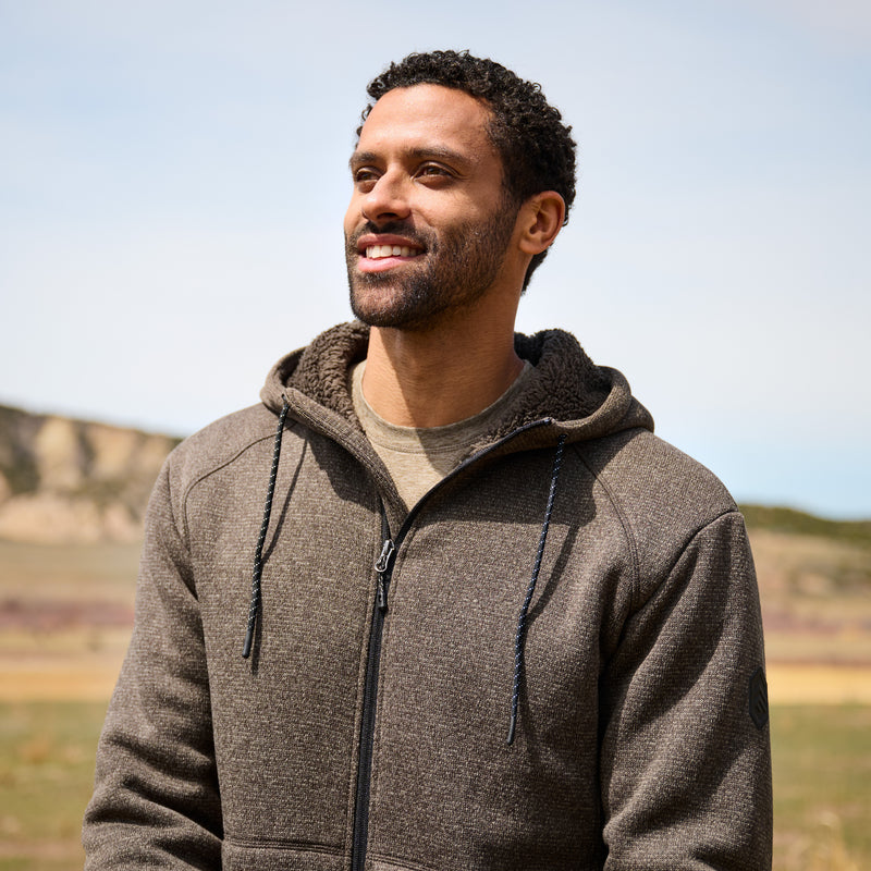 Man wearing a brown hoodie standing outdoors with a clear sky background