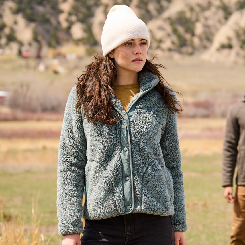 Woman wearing a green fleece jacket and white beanie outdoors with mountains in the background