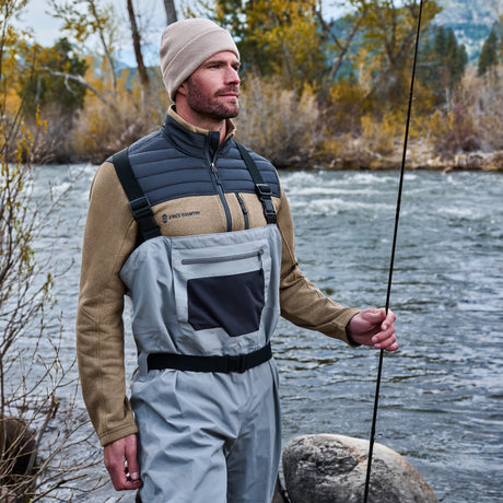 A man by a river in waders and a beige beanie holds a fishing rod, wearing the Men’s Frore Knit Fleece Jacket from Free Country, surrounded by autumn trees and rocks.