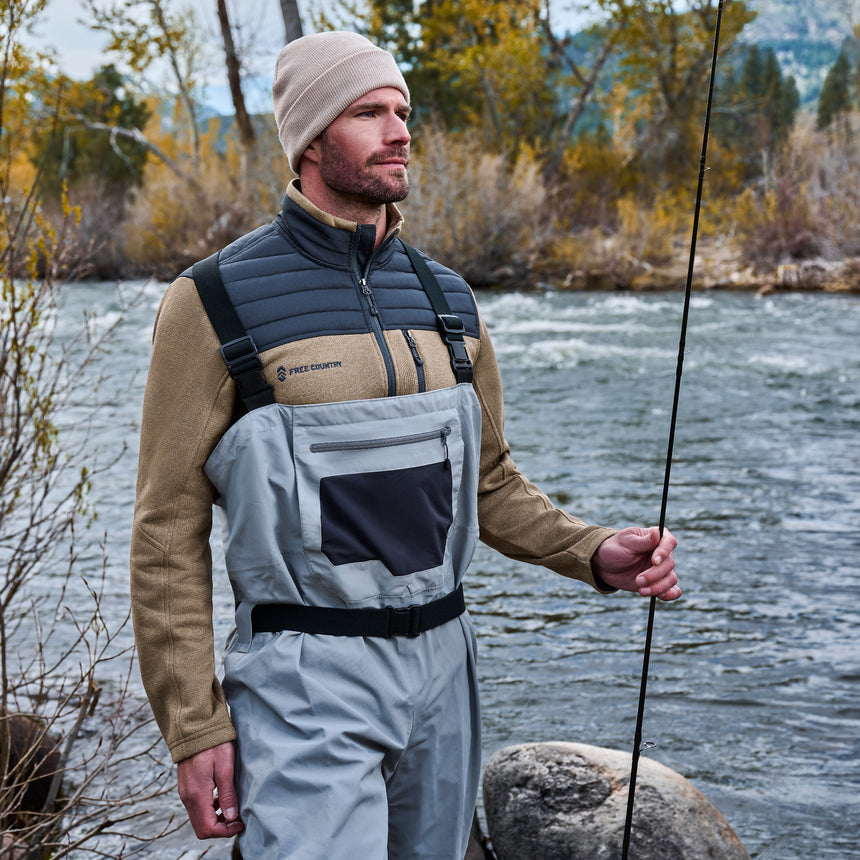A man by a river in waders and a beige beanie holds a fishing rod, wearing the Men’s Frore Knit Fleece Jacket from Free Country, surrounded by autumn trees and rocks.