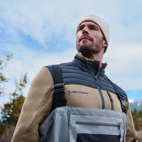 A man in outdoor gear, including a beige beanie and the Free Country Men’s Frore Knit Fleece Jacket, gazes into the distance with trees and a cloudy sky in the background.