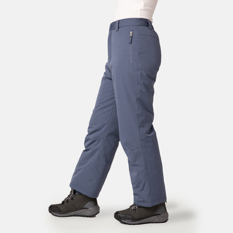 A person is shown from the waist down wearing Free Country Kids' Padded Ski Pant in blue, featuring a zipper pocket and built-in snow gaiters, with black hiking boots, walking on a plain light background—perfect for outdoor activities.