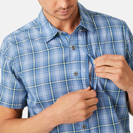 A man wearing the Free Country Men's Excursion Short Sleeve Poplin Shirt in blue plaid opens a hidden zippered chest pocket, showing the mesh lining inside. The man's face is partially out of frame.