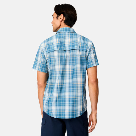 A man stands with his back to the camera, wearing a light blue and white Free Country Men's Excursion Short Sleeve Poplin Shirt and dark blue pants against a plain white background.