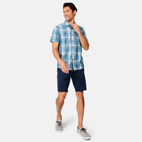 A man smiles while wearing the Free Country Men's Excursion Short Sleeve Poplin Shirt in blue plaid, navy shorts, and light gray sneakers, standing against a plain white background.