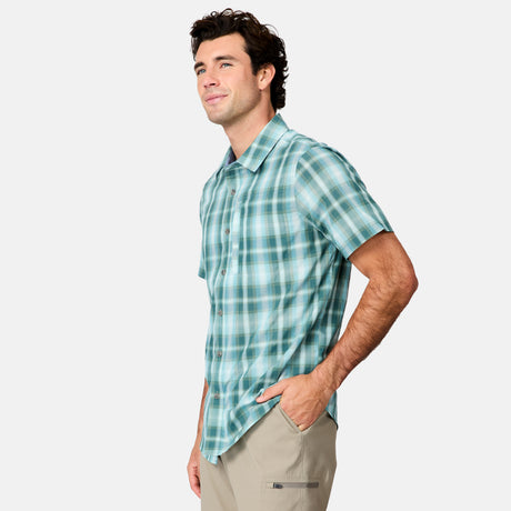 A man wears the Free Country Men's Excursion Short Sleeve Poplin Shirt in teal and white plaid with quick dry fabric, paired with light beige pants. He stands against a plain light background, smiling with one hand in his pocket.