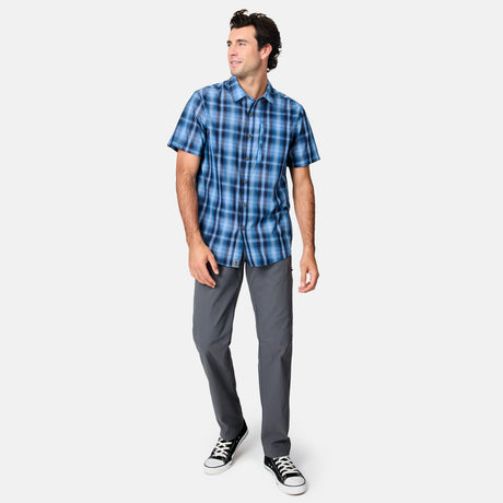 A man stands against a plain white background, wearing the Free Country Men's Excursion Short Sleeve Poplin Shirt in blue plaid, paired with gray pants and black-and-white sneakers. He is looking to the side and smiling.