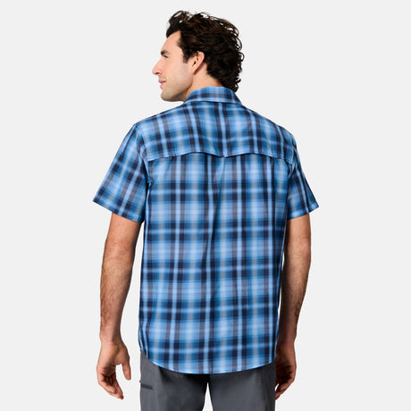 A man with dark hair wears the Free Country Men's Excursion Short Sleeve Poplin Shirt in blue plaid and gray pants, standing with his back to the camera against a plain light background.