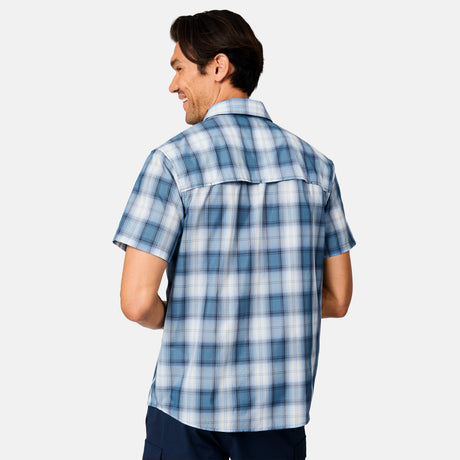 A man with dark hair is seen from behind wearing the Free Country Men's Excursion Short Sleeve Poplin Shirt in blue and white plaid, made from quick dry fabric, along with dark pants, standing against a plain light background.