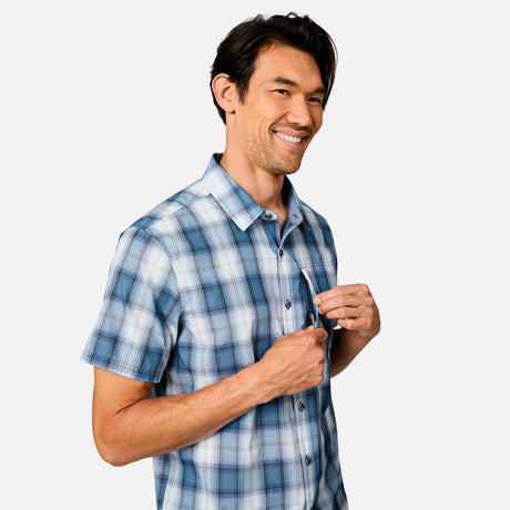 A man wearing the Free Country Men's Excursion Short Sleeve Poplin Shirt smiles while buttoning the quick-dry, moisture-wicking shirt against a plain light background.