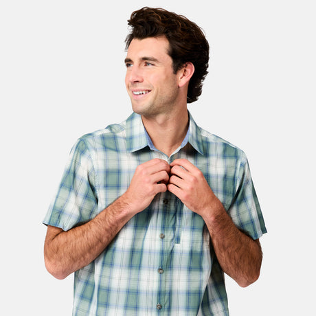 A brown-haired man smiles and looks to the side while buttoning the Free Country Men's Excursion Short Sleeve Poplin Shirt in blue and green plaid, standing against a plain light gray background.