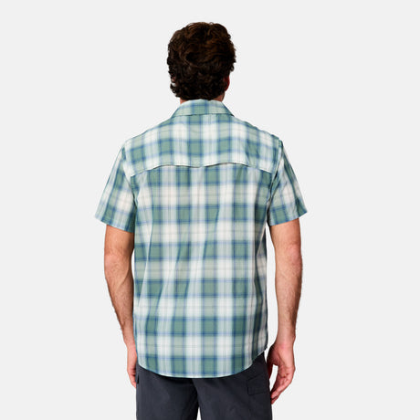 A man with short brown hair stands with his back to the camera, wearing a moisture-wicking Free Country Men's Excursion Short Sleeve Poplin Shirt in light blue and green plaid, with dark gray shorts, against a plain white background.