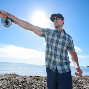 A man in the Free Country Men's Excursion Short Sleeve Poplin Shirt, sunglasses, and a cap stands on a rocky shoreline fishing with a reel under a bright sunny sky by the water.