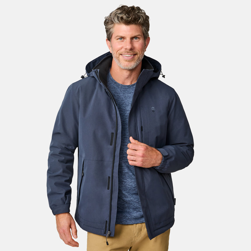 [Kolby is 6’2” wearing size M.] Men’s Free Country navy zip-front jacket with hood and high collar, featuring vertical chest zipper pocket and side hand pockets—worn over light brown pants for an outdoors-ready, insulated look.