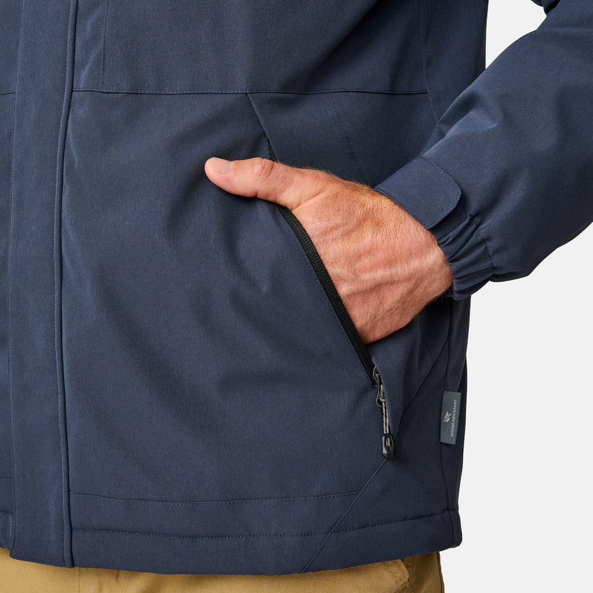 [Kolby is 6’2” wearing size M.] Men’s Free Country navy zip-front jacket with hood and high collar, featuring vertical chest zipper pocket and side hand pockets—worn over light brown pants for an outdoors-ready, insulated look.